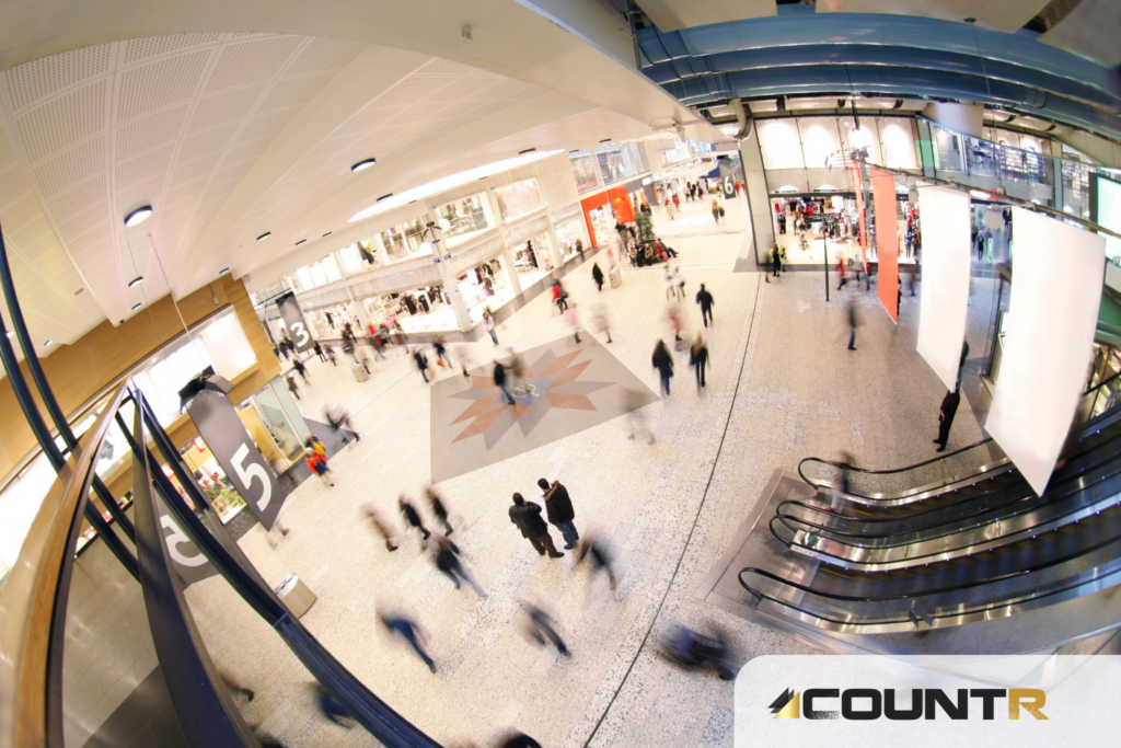 Wide-angle view of a busy shopping mall entrance with people walking and escalators visible 
